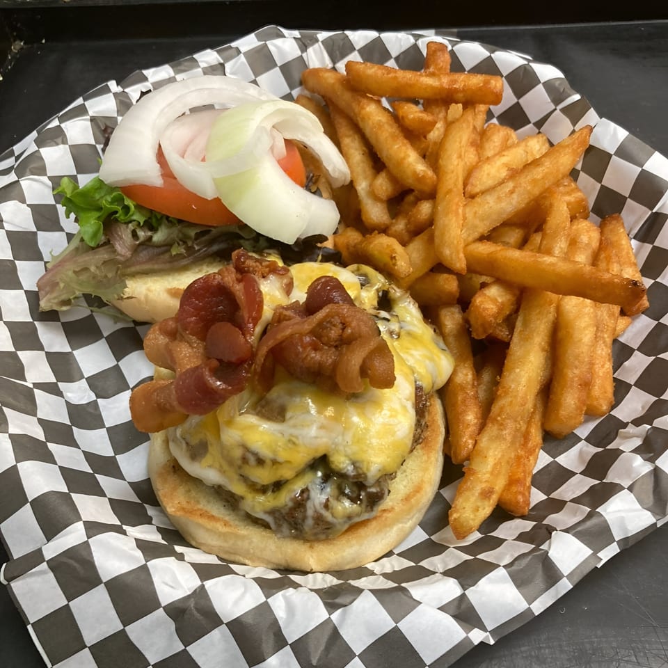 Bacon cheeseburger with lettuce, tomato, onion, and a side of fries in a checkered basket.