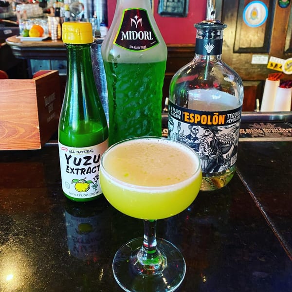 A vibrant green cocktail in a coupe glass, with bottles of Midori, Yuzu Extract, and Espolon Tequila.