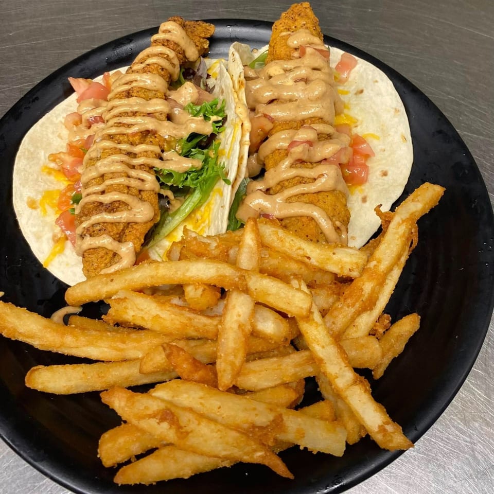 Two crispy fish tacos with sauce, lettuce, and tomatoes, served with a side of golden fries.