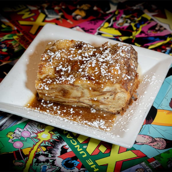 Bread pudding with powdered sugar and caramel sauce on a white plate, atop comic books.