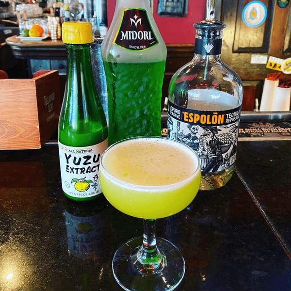 A bright green cocktail in a coupe glass sits on a bar next to bottles of Midori, Yuzu Extract, and tequila.