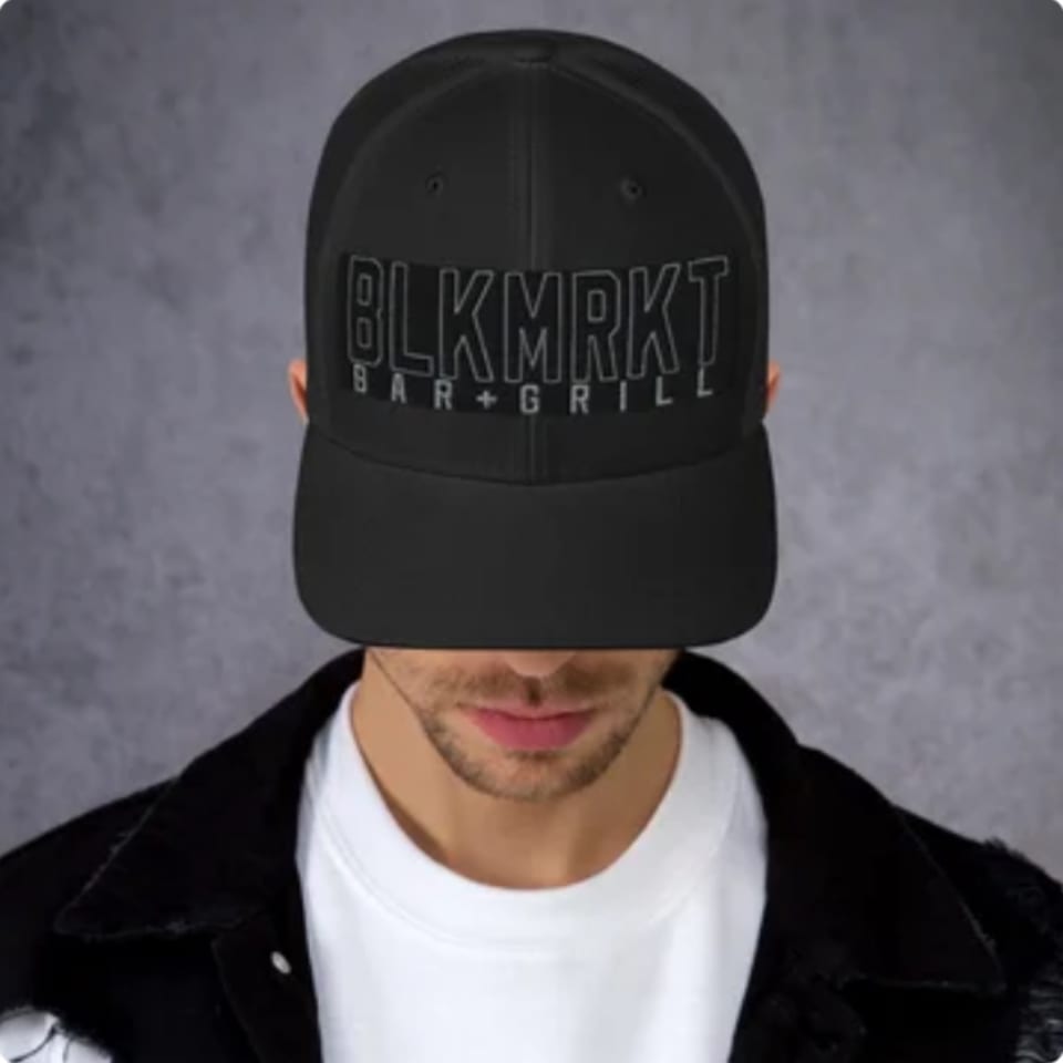 A person wearing a black Black Market Bar + Grill baseball cap and a black jacket.