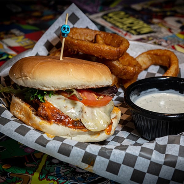 A Black Market Bar and Grill burger with melted cheese, lettuce, and tomato, served with onion rings and dipping sauce.