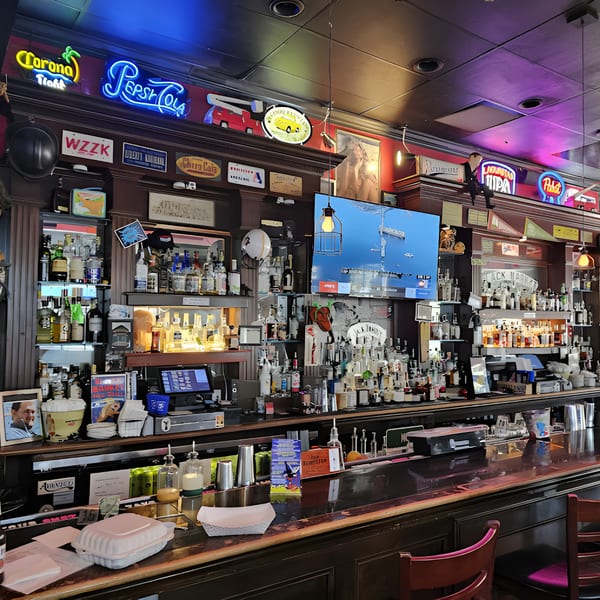 A well-stocked bar at Black Market Bar and Grill, with neon signs and a TV displaying a boat scene.
