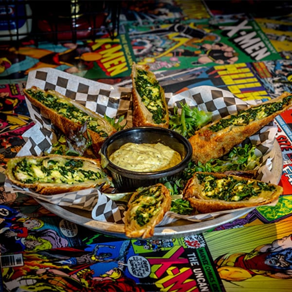 Spinach and artichoke egg rolls with dipping sauce on a comic book themed table at Black Market Bar and Grill.