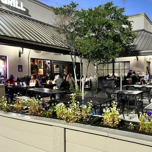 Patrons seated at outdoor tables on the patio of Black Market Bar and Grill, with trees and planters.