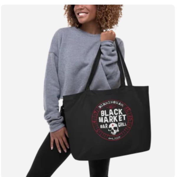 Smiling woman in a gray sweatshirt holding a Black Market Bar & Grill logo tote bag.